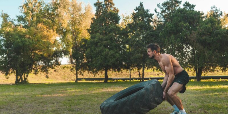 Athletic man flipping large tire during outdoor workout in sunny park setting.