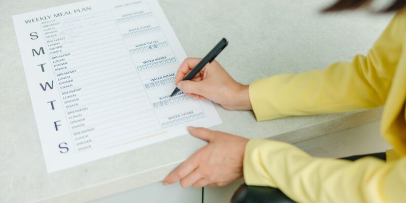 Woman writing on a weekly meal plan checklist indoors, focusing on diet organization.