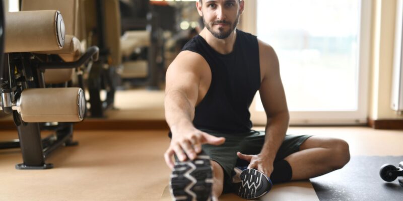 A man in gym clothes stretches on the floor in a modern fitness center.