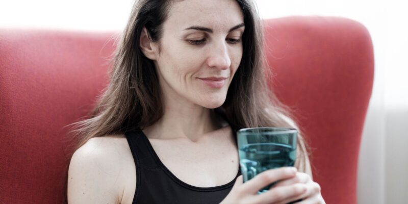 A relaxed woman in black top holding a glass of water and smiling, sitting indoors.