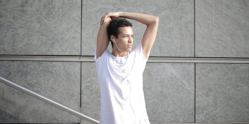 Calm pensive African American sportsman in earphones and sportswear looking away and thinking while stretching arms and listening to music with pleasure on street against gray exterior of contemporary buildings